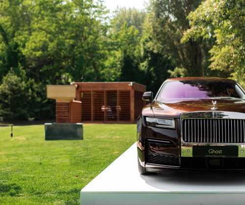 A luxurious black Rolls-Royce parked on a white platform with a wooden cabin and greenery in the background.